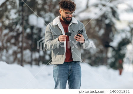 Man with curly hair wearing a gray jacket and glasses is using a smartphone while holding a credit card outdoors in a snowy environment with trees in the background Man with curly hair wearing a gray jacket and glasses is using a smartphone while holding a credit card outdoors in a snowy environment with trees in the background 140031851
