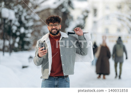 Young man with curly hair wearing a gray jacket and glasses is holding a shopping bag while using a smartphone in a snowy urban setting with blurred pedestrians Young man with curly hair wearing a gray jacket and glasses is holding a shopping bag while using a smartphone in a snowy urban setting with blurred pedestrians 140031858