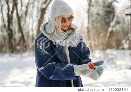 Young woman wearing winter clothing, gray hat and scarf, holds a small orange object while standing in a snowy forest with trees in the background Young woman wearing winter clothing, gray hat and scarf, holds a small orange object while standing in a snowy forest with trees in the background 140032104