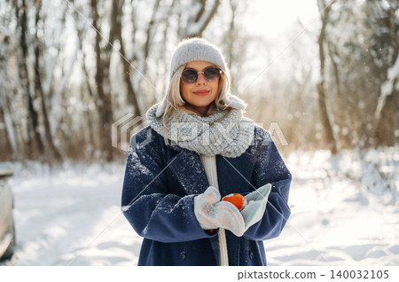 Young woman in winter attire holding oranges, wearing sunglasses and a knitted scarf, standing in a snowy forest with trees in the background Young woman in winter attire holding oranges, wearing sunglasses and a knitted scarf, standing in a snowy forest with trees in the background 140032105