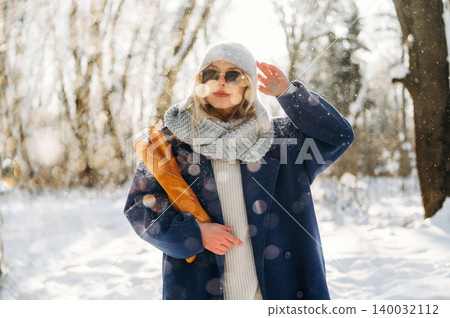 Young woman in winter attire wearing sunglasses holds a baguette while standing in a snowy landscape with trees and sunlight filtering through Young woman in winter attire wearing sunglasses holds a baguette while standing in a snowy landscape with trees and sunlight filtering through 140032112