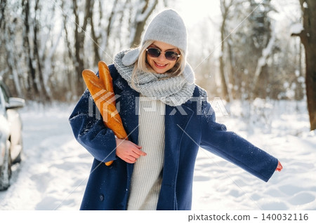Young woman in winter attire holds fresh baguettes while walking through a snowy landscape, trees and a parked car visible in the background 140032116