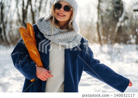 Young woman in winter attire wearing sunglasses holds fresh baguettes while standing in a snowy landscape with trees in the background Young woman in winter attire wearing sunglasses holds fresh baguettes while standing in a snowy landscape with trees in the background 140032117