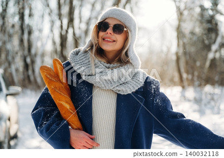 Woman in winter attire wearing sunglasses holds a fresh baguette while standing outdoors in a snowy landscape with trees in the background Woman in winter attire wearing sunglasses holds a fresh baguette while standing outdoors in a snowy landscape with trees in the background 140032118