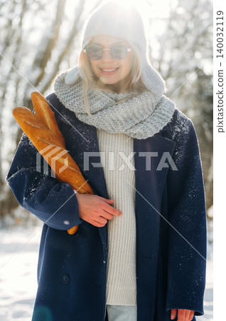 Young woman wearing sunglasses and a warm coat holds fresh baguettes while standing in a snowy outdoor setting with trees in the background Young woman wearing sunglasses and a warm coat holds fresh baguettes while standing in a snowy outdoor setting with trees in the background 140032119
