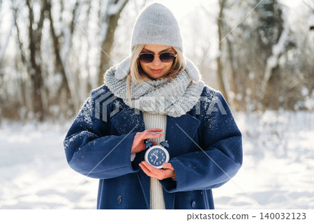 Young woman in winter attire holding a vintage alarm clock while standing in a snowy forest, wearing sunglasses and a knitted scarf, with snow-covered trees in the background Young woman in winter attire holding a vintage alarm clock while standing in a snowy forest, wearing sunglasses and a knitted scarf, with snow-covered trees in the background 140032123