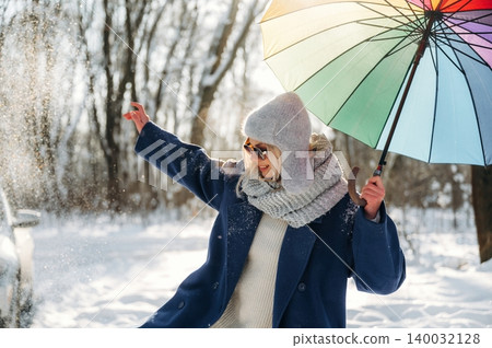 Woman in winter attire joyfully plays with a colorful umbrella while standing in a snowy landscape with trees in the background and sunlight filtering through branches Woman in winter attire joyfully plays with a colorful umbrella while standing in a snowy landscape with trees in the background and sunlight filtering through branches 140032128