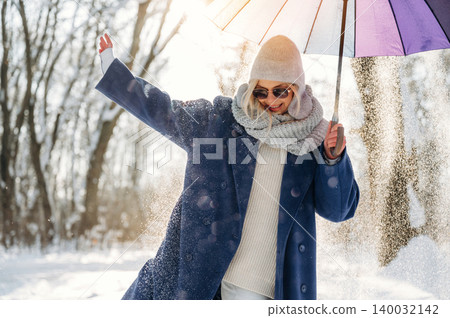 Woman in winter attire joyfully holding a colorful umbrella while standing in a snowy forest, sunlight filtering through trees creating a bright atmosphere Woman in winter attire joyfully holding a colorful umbrella while standing in a snowy forest, sunlight filtering through trees creating a bright atmosphere 140032142