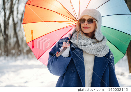 Woman in winter attire holding a colorful umbrella stands in a snowy landscape, wearing sunglasses and a cozy scarf, surrounded by trees in a winter setting Woman in winter attire holding a colorful umbrella stands in a snowy landscape, wearing sunglasses and a cozy scarf, surrounded by trees in a winter setting 140032143