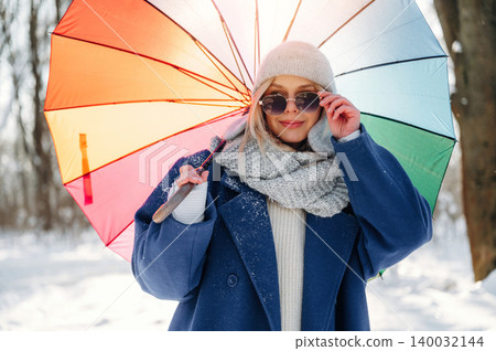 Young woman wearing sunglasses and a cozy scarf holds a colorful umbrella while standing in a snowy landscape with trees in the background 140032144