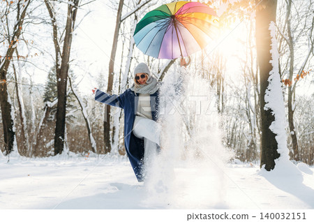 Woman in blue coat and gray scarf joyfully plays in snow while holding a colorful umbrella in a winter forest setting with sunlight filtering through trees 140032151