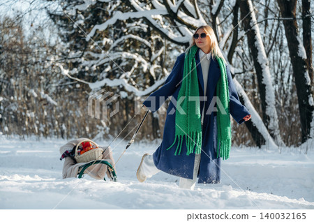 Woman in blue coat and green scarf walks through snowy landscape, pulling a sled with a child and winter trees in the background 140032165