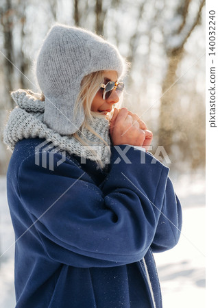 Woman wearing gray knitted hat and sunglasses holds hands together while standing in snowy forest, sunlight filtering through trees in winter landscape Woman wearing gray knitted hat and sunglasses holds hands together while standing in snowy forest, sunlight filtering through trees in winter landscape 140032240