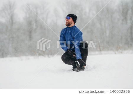 Male athlete in blue jacket and black pants crouches in snow-covered field during winter snowfall, trees blurred in the background 140032896