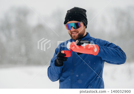 Male hiker wearing sunglasses and a black beanie pours hot beverage from a red thermos while standing in a snowy landscape with trees in the background Male hiker wearing sunglasses and a black beanie pours hot beverage from a red thermos while standing in a snowy landscape with trees in the background 140032900