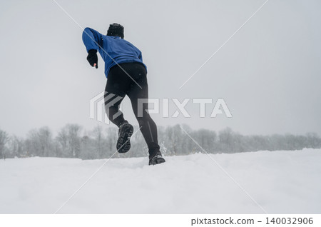Male runner in blue jacket and black leggings sprinting through deep snow in a winter landscape with trees and overcast sky in the background Male runner in blue jacket and black leggings sprinting through deep snow in a winter landscape with trees and overcast sky in the background 140032906