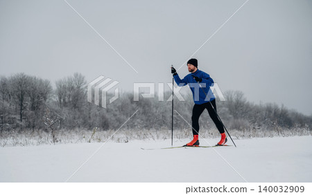 Male cross-country skier in blue jacket and black pants glides through snowy landscape with trees covered in frost under overcast sky 140032909