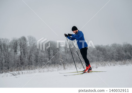 Male cross-country skier in blue jacket and black pants glides over snowy landscape with trees covered in frost in the background during winter season Male cross-country skier in blue jacket and black pants glides over snowy landscape with trees covered in frost in the background during winter season 140032911