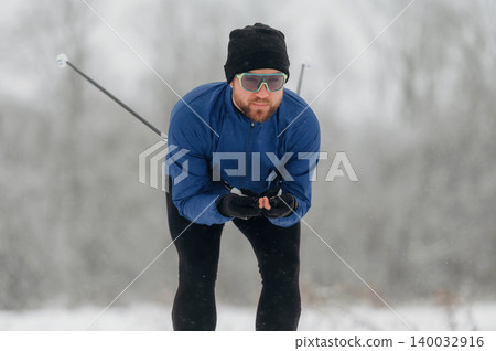 Male cross-country skier in blue jacket and black pants gliding through snowy landscape, wearing sunglasses and gloves, with trees blurred in the background Male cross-country skier in blue jacket and black pants gliding through snowy landscape, wearing sunglasses and gloves, with trees blurred in the background 140032916
