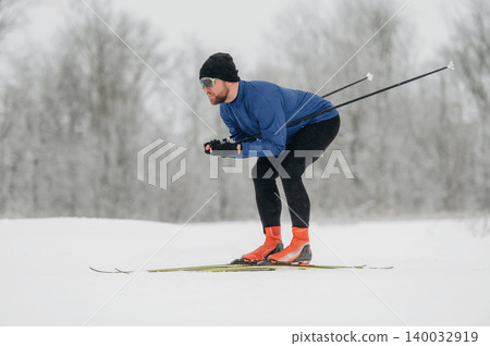 Male cross-country skier in blue long-sleeve shirt and black pants gliding over snow-covered terrain with trees in the background during winter season Male cross-country skier in blue long-sleeve shirt and black pants gliding over snow-covered terrain with trees in the background during winter season 140032919
