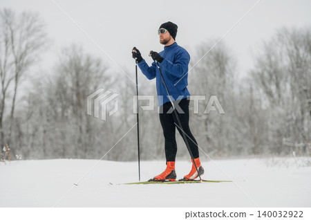 Male cross-country skier in blue jacket and black pants stands on snow-covered trail, holding ski poles with trees in the background during winter season Male cross-country skier in blue jacket and black pants stands on snow-covered trail, holding ski poles with trees in the background during winter season 140032922