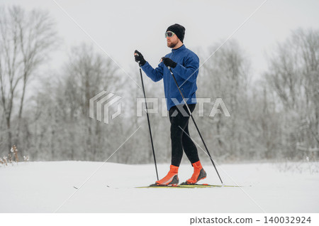 Male cross-country skier in blue jacket and black leggings glides over snow-covered landscape with trees in the background during winter season 140032924