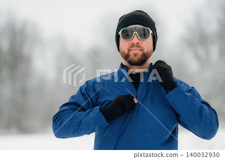 Man in blue jacket and sunglasses adjusts zipper while standing in snowy landscape with blurred trees in the background, winter sports attire visible Man in blue jacket and sunglasses adjusts zipper while standing in snowy landscape with blurred trees in the background, winter sports attire visible 140032930