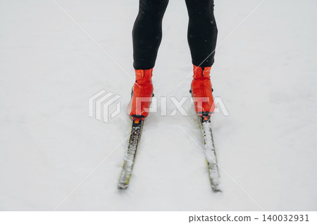 Individual wearing black leggings and bright orange ski boots stands on snow-covered ground with cross-country skis positioned parallel in a winter landscape Individual wearing black leggings and bright orange ski boots stands on snow-covered ground with cross-country skis positioned parallel in a winter landscape 140032931