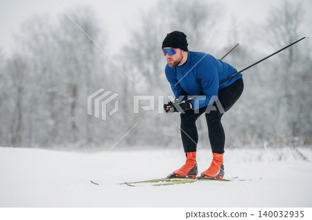 Male cross-country skier in blue jacket and black tights gliding on snow-covered trail, winter landscape with trees and overcast sky in the background Male cross-country skier in blue jacket and black tights gliding on snow-covered trail, winter landscape with trees and overcast sky in the background 140032935