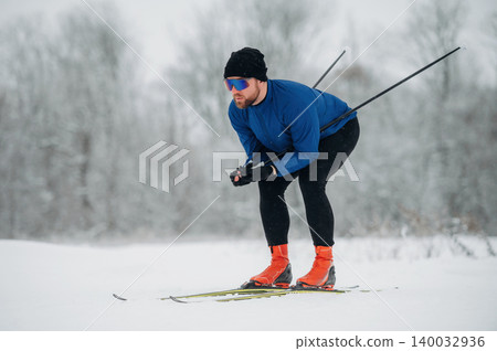 Male cross-country skier in blue long-sleeve shirt and black pants glides over snow-covered terrain with trees in the background during winter sports activity Male cross-country skier in blue long-sleeve shirt and black pants glides over snow-covered terrain with trees in the background during winter sports activity 140032936