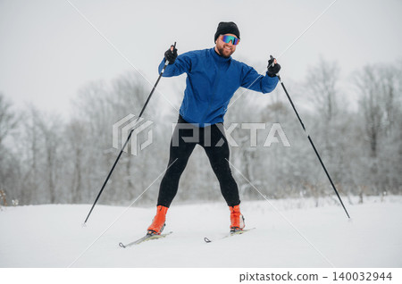 Male cross-country skier in blue jacket and black pants glides through snowy landscape, wearing sunglasses and ski poles, with trees in the background Male cross-country skier in blue jacket and black pants glides through snowy landscape, wearing sunglasses and ski poles, with trees in the background 140032944