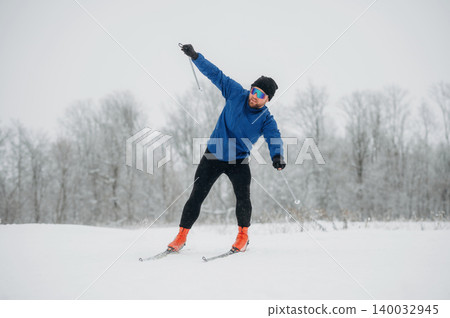Male skier in blue jacket and black pants glides on snow-covered trail with trees in the background during winter sports activity in a cold environment Male skier in blue jacket and black pants glides on snow-covered trail with trees in the background during winter sports activity in a cold environment 140032945