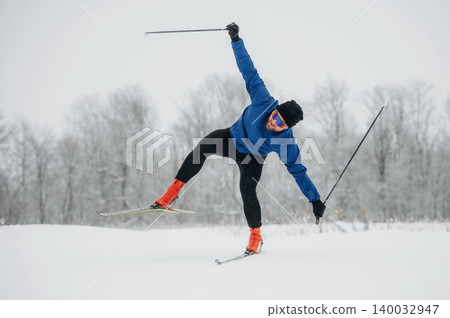 Male cross-country skier in blue jacket and black pants performs a dynamic maneuver on snow-covered trail with trees in the background during winter season 140032947