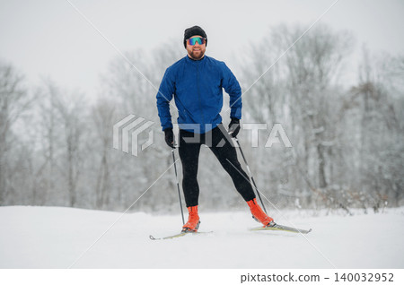 Male cross-country skier in blue jacket and black pants stands on snow-covered trail, wearing sunglasses and ski poles, surrounded by winter trees in a snowy landscape 140032952