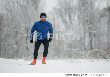 Male cross-country skier in blue jacket and black pants glides through snowy landscape, wearing sunglasses and bright orange ski boots amid winter trees 140032953