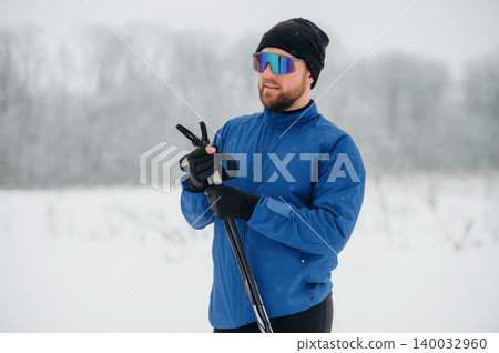 Male skier in blue jacket and sunglasses holds ski poles while standing in snowy landscape with blurred trees in the background during winter season Male skier in blue jacket and sunglasses holds ski poles while standing in snowy landscape with blurred trees in the background during winter season 140032960