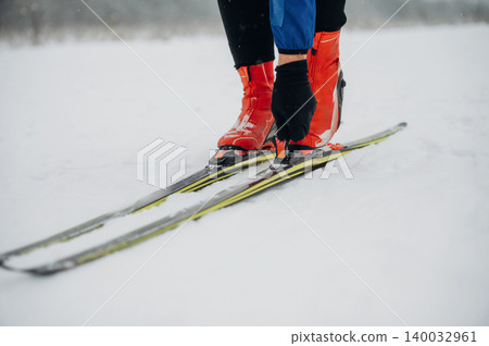 Skier wearing red boots adjusts bindings on cross-country skis while standing on snow-covered terrain in a winter landscape with blurred trees in the background Skier wearing red boots adjusts bindings on cross-country skis while standing on snow-covered terrain in a winter landscape with blurred trees in the background 140032961