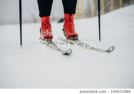 Skier wearing bright red boots stands on snow-covered slope with ski poles, showcasing winter sports equipment and snowy landscape in the background Skier wearing bright red boots stands on snow-covered slope with ski poles, showcasing winter sports equipment and snowy landscape in the background 140032966
