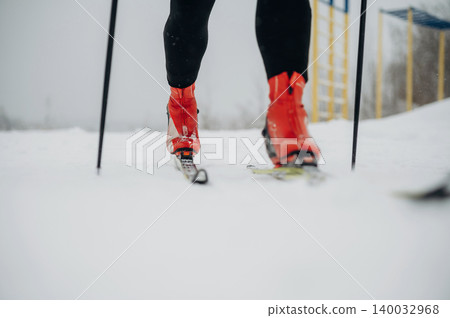 Skier wearing red boots and black pants glides on snow-covered terrain with ski poles, yellow structures visible in the background under overcast sky Skier wearing red boots and black pants glides on snow-covered terrain with ski poles, yellow structures visible in the background under overcast sky 140032968