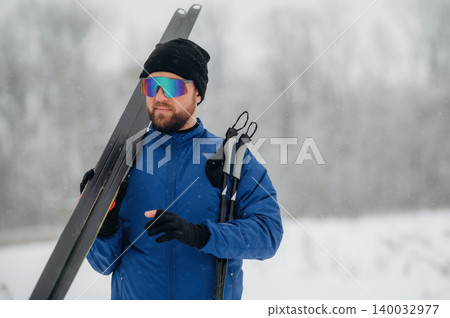 Male skier wearing blue jacket and sunglasses holds skis and poles in snowy landscape with blurred trees in the background during winter season Male skier wearing blue jacket and sunglasses holds skis and poles in snowy landscape with blurred trees in the background during winter season 140032977