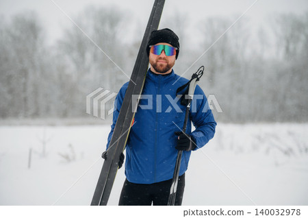 Male skier wearing blue jacket and sunglasses holds skis and poles in snowy landscape with trees and falling snowflakes in the background Male skier wearing blue jacket and sunglasses holds skis and poles in snowy landscape with trees and falling snowflakes in the background 140032978