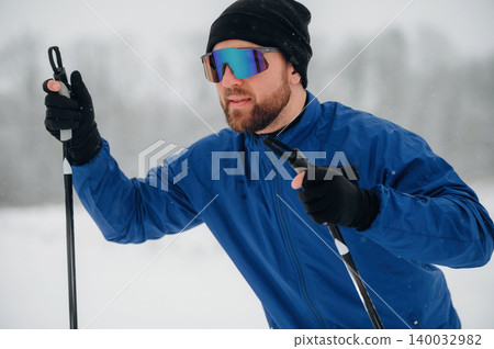 Male skier wearing blue jacket and sunglasses holds ski poles while preparing to ski in a snowy landscape with blurred trees in the background Male skier wearing blue jacket and sunglasses holds ski poles while preparing to ski in a snowy landscape with blurred trees in the background 140032982