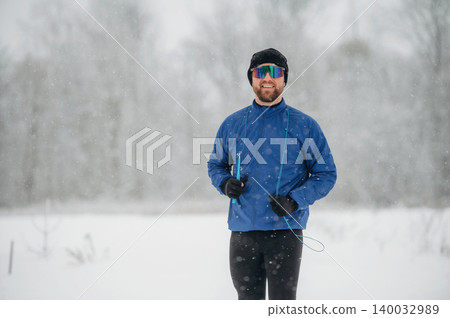 Male athlete wearing blue jacket and sunglasses stands in snowy landscape, snowflakes falling around him, winter trees blurred in the background Male athlete wearing blue jacket and sunglasses stands in snowy landscape, snowflakes falling around him, winter trees blurred in the background 140032989