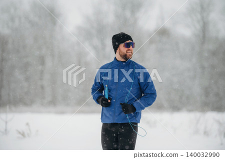 Male runner dressed in blue jacket and black pants jogs through snowy landscape wearing sunglasses and headphones during winter weather with trees in the background Male runner dressed in blue jacket and black pants jogs through snowy landscape wearing sunglasses and headphones during winter weather with trees in the background 140032990