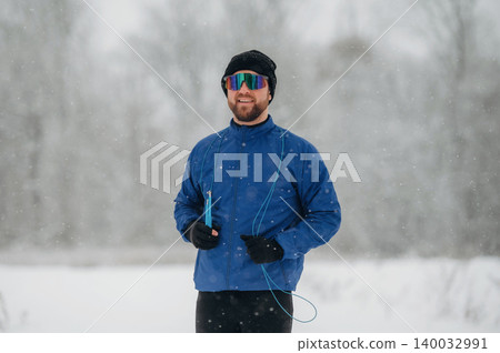 Male athlete wearing blue jacket and sunglasses stands in snowy landscape, holding a jump rope with snowflakes falling around him in a winter outdoor setting Male athlete wearing blue jacket and sunglasses stands in snowy landscape, holding a jump rope with snowflakes falling around him in a winter outdoor setting 140032991