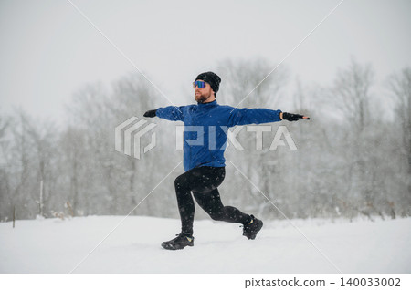 Male athlete performing a lunge exercise in snowy outdoor setting, wearing blue jacket, black pants, gloves, and sunglasses, with trees covered in snow in the background Male athlete performing a lunge exercise in snowy outdoor setting, wearing blue jacket, black pants, gloves, and sunglasses, with trees covered in snow in the background 140033002