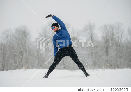 Male athlete stretching in snow-covered landscape during winter, wearing blue jacket and black leggings, with trees and snowfall in the background 140033005