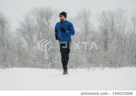 Male runner dressed in blue long-sleeve shirt and black leggings jogs through snowy landscape with trees in the background during winter weather 140033006