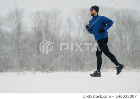Male runner in blue long-sleeve shirt and black leggings jogs through snowy landscape with trees in the background during winter weather conditions 140033007