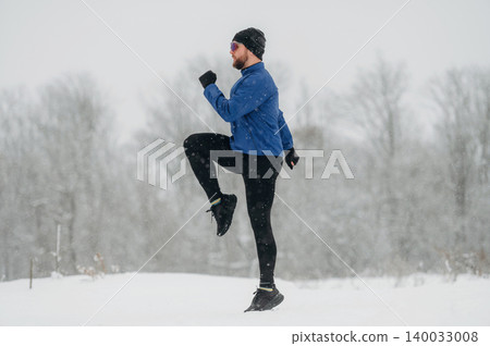 Male runner in blue jacket and black leggings exercises in snowy landscape, snowflakes falling, trees blurred in the background, winter fitness activity 140033008